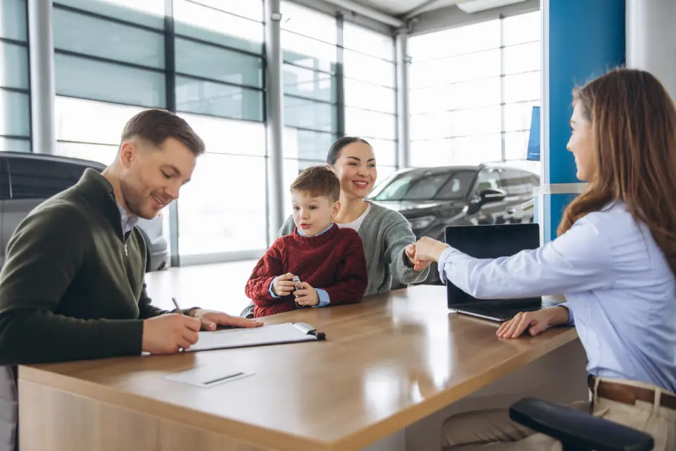 Family at a car dealership: father signs paperwork at a desk while mother holds their child, as the salesperson shakes hands with them.